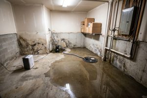 Wide view of a water-damaged basement with standing water, moisture damage on drywall, and a sump pump in operation.